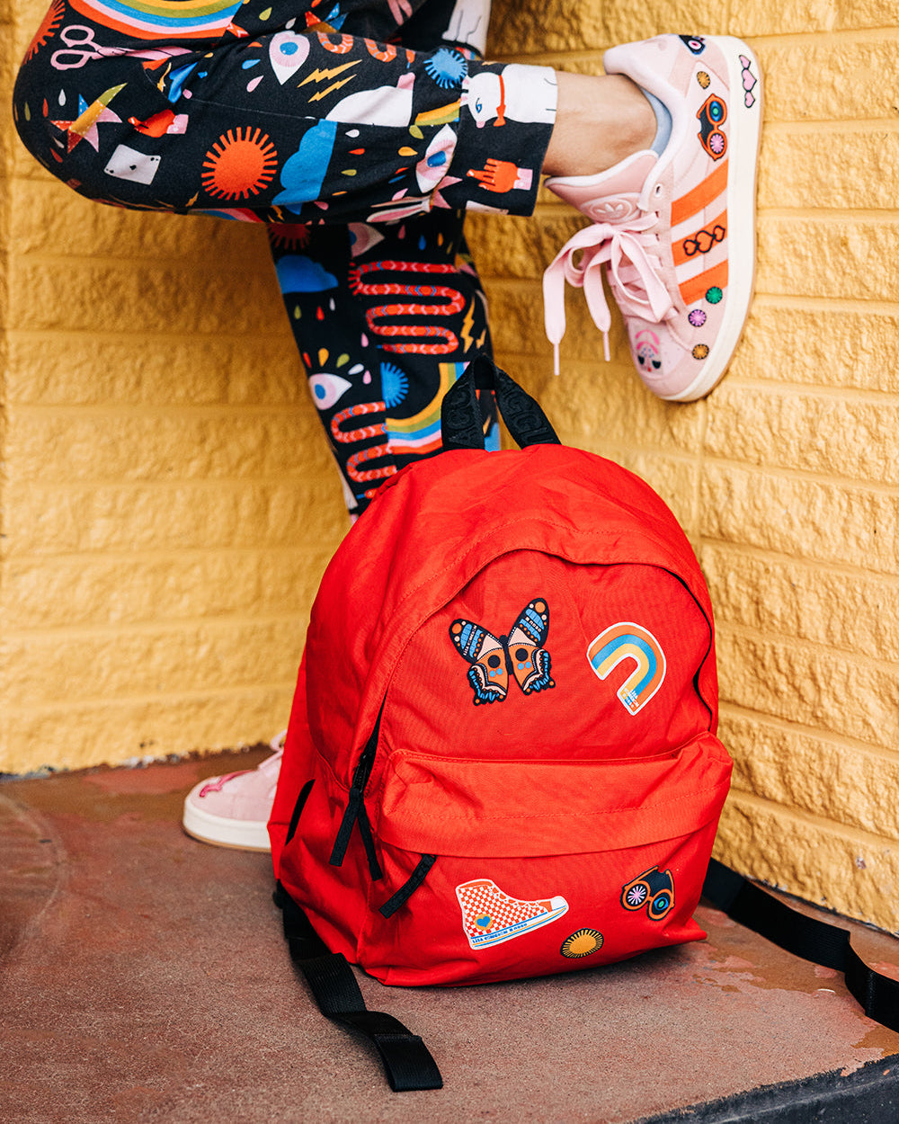 Person wearing colorful pants and shoes with a red backpack against a yellow textured wall featuring NoSo Patches.