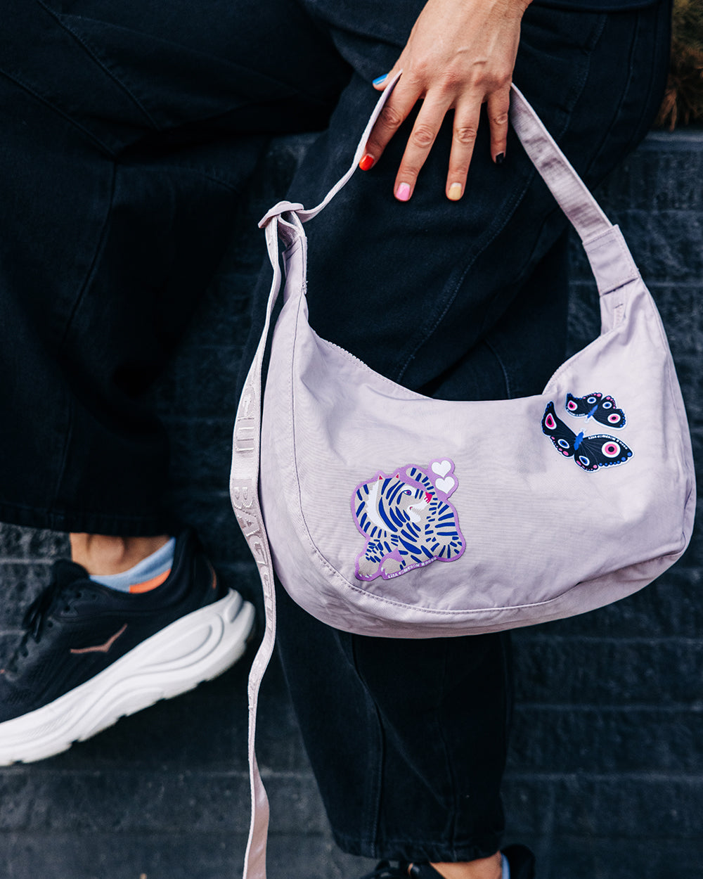Person holding a light purple handbag with decorative NoSo Patches, wearing black pants and sneakers.