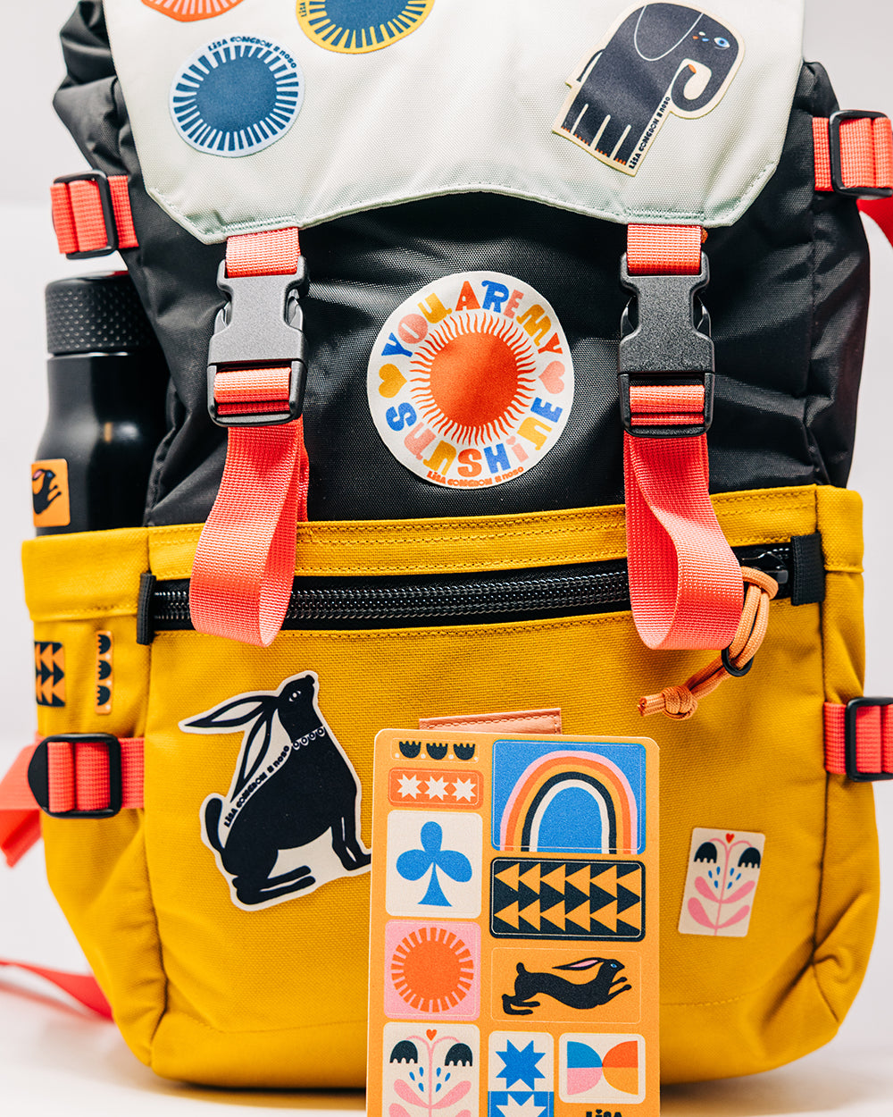 Colorful backpack with various Lisa Cogdon NoSo Patches and a water bottle on a white background