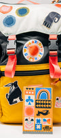 Colorful backpack with various Lisa Cogdon NoSo Patches and a water bottle on a white background