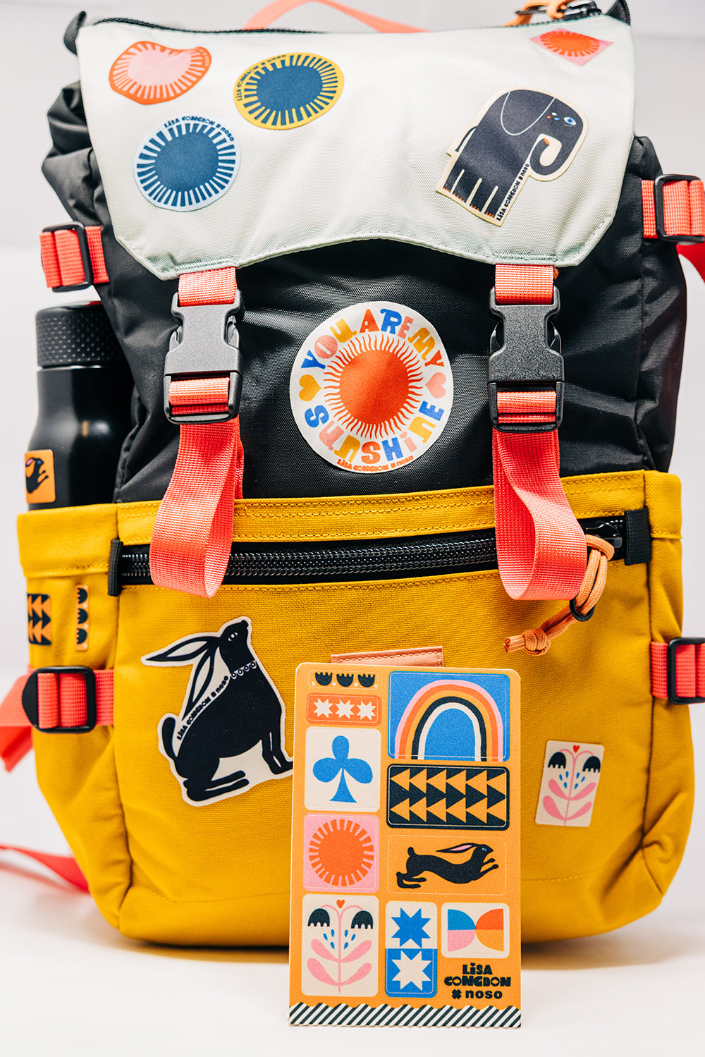 Colorful backpack with various Lisa Cogdon NoSo Patches and a water bottle on a white background